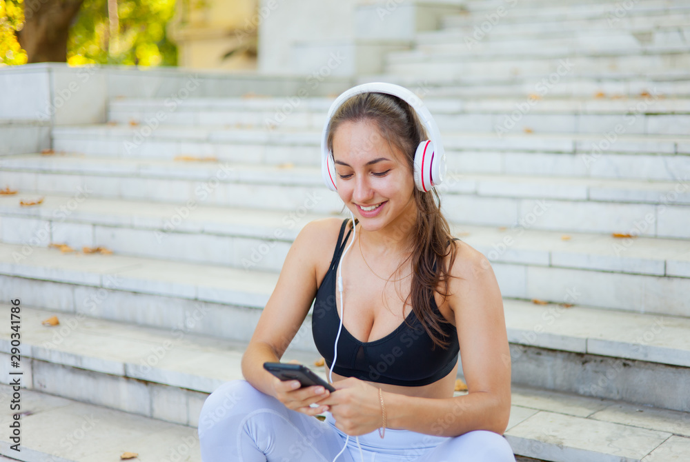Concept of rest after training. Young happy sport woman in sportswear use smartphone and listens to music in headphones while sitting on stairs on bright sunny day Concept of rest after training. Young happy sport woman in sportswear use smartphone and listens to music in headphones while sitting on stairs on bright sunny day