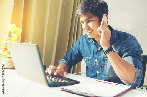 Attractive asian guy talking on phone while using laptop at workplace.