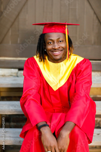 Smiling happy African-American Teen Teenager male Man outside against a brown wall in his red and gold graduation gown