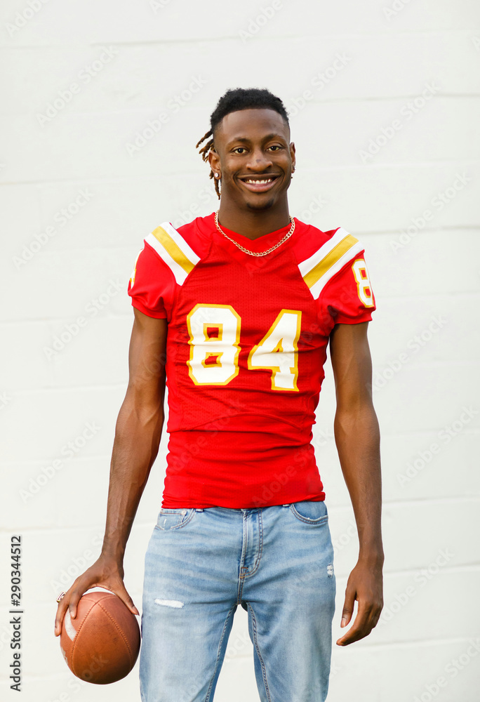 happy smiling young Handsome African American Black Male outside wearing a  red and gold football jersey and holding a football. he is wearing jeans  and he is a high school student-athlete Stock