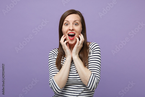 Amazed surprised young woman in casual striped clothes posing isolated on violet purple background in studio. People lifestyle concept. Mock up copy space. Keeping mouth open, putting hands on face.