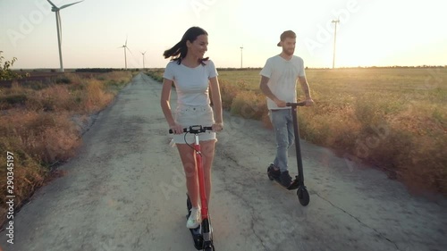 A guy and a young girl at sunset ride on electric scooters on the background of wind power generators. Alternative energy.