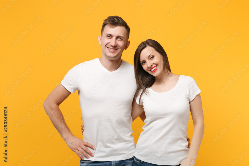 Pretty cheerful young couple two friends guy girl in white empty blank design t-shirts posing isolated on yellow orange wall background studio portrait. People lifestyle concept. Mock up copy space. Pretty cheerful young couple two friends guy girl in white empty blank design t-shirts posing isolated on yellow orange wall background studio portrait. People lifestyle concept. Mock up copy space.