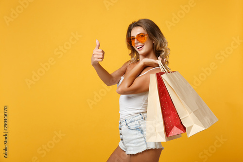Side view of smiling woman girl in eyeglasses posing isolated on yellow background. People lifestyle concept. Mock up copy space. Holding package bag with purchases after shopping, showing OK gesture.