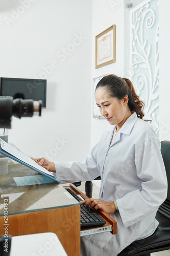 Asian female doctor in white coat sitting at her worklace and typing on computer keyboard at office