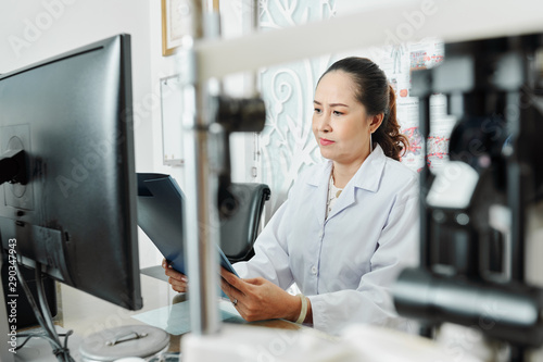 Asian mature woman sitting at the table in front of computer with folder and reading a medical card at office