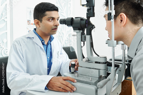 Indian optometrist in white coat looking through the special medical equipment and examining his patient's eyesight at hospital