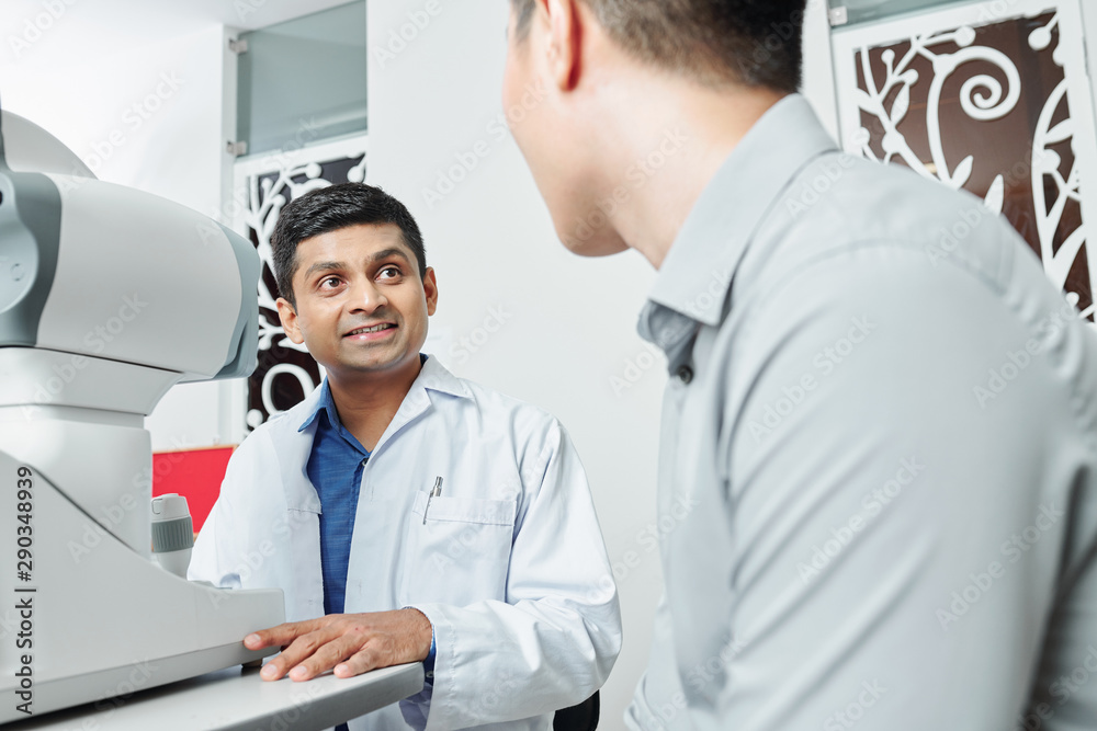Indian doctor in white coat sitting at the table in front of medical equipment and talking to the patient during his visit at office Indian doctor in white coat sitting at the table in front of medical equipment and talking to the patient during his visit at office