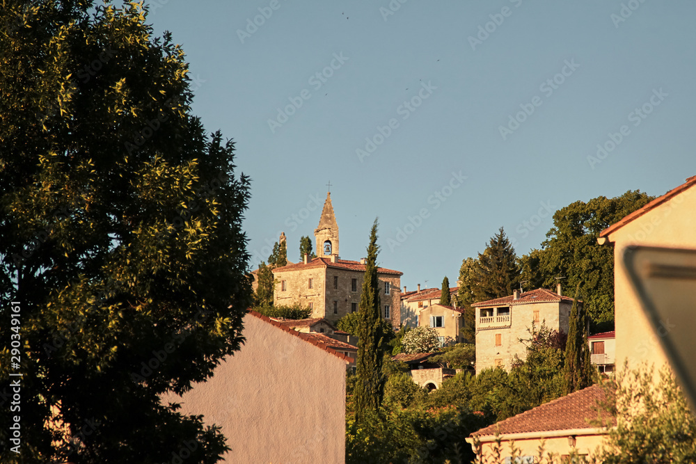 View of  the old town of Barjac, southern France. Green trees, roofs of buildings and stone church with a bell tower on a hill at sunset light