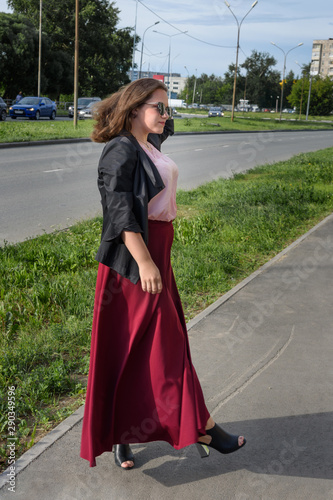 Brunette girl walking on the street in city. Girl in black jacket and red skirt with long hair. Plus size model © Elena Odareeva