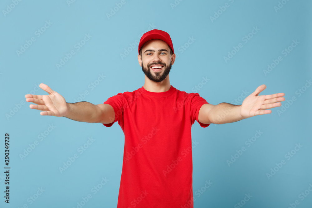 Naklejka premium Delivery man in red uniform workwear isolated on blue wall background, studio portrait. Professional male employee in cap t-shirt print working as courier dealer. Service concept. Mock up copy space.