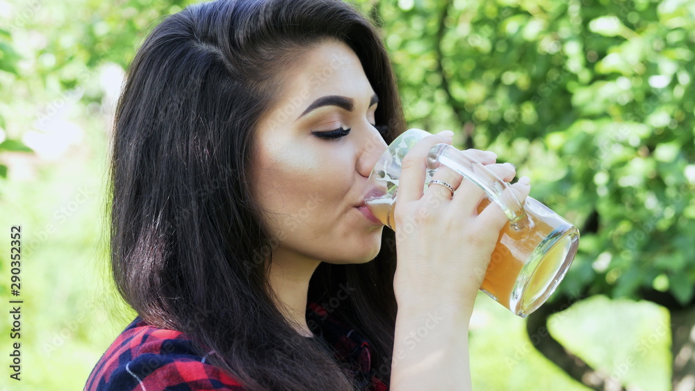 Side portrait of young happy beautiful woman which drinks beer from glass on picnic then looking at the camera and smiling, quenches thirst Side portrait of young happy beautiful woman which drinks beer from glass on picnic then looking at the camera and smiling, quenches thirst