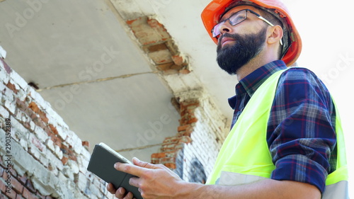 An engineer builder in a helmet is calculating the building demolition plan and looking at the construction project on a tablet, the ruins in the background