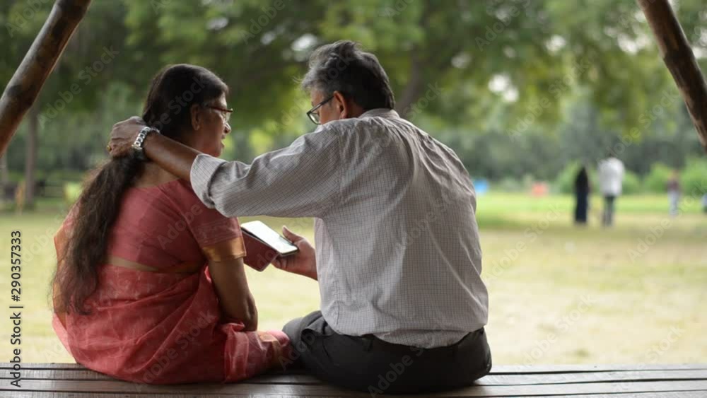 Senior citizen Indian man reads from mobile and puts hand over shoulder shot from behind on his wife while sitting in a park in Delhi, India Senior citizen Indian man reads from mobile and puts hand over shoulder shot from behind on his wife while sitting in a park in Delhi, India