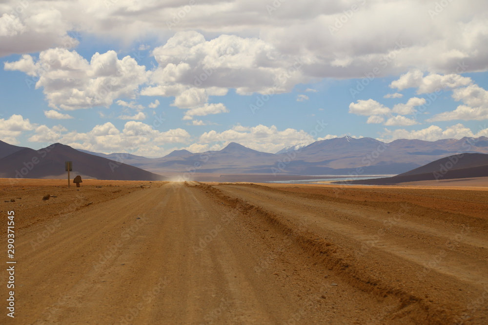 Valley with wide dusty road with light brown soil. Cloud sky and mountains in the horizon. A off road vehicle running and raising dusty. Bolivia highlands desert. Valley with wide dusty road with light brown soil. Cloud sky and mountains in the horizon. A off road vehicle running and raising dusty. Bolivia highlands desert.