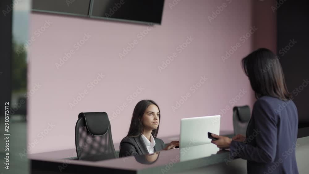 Businesswoman talking with young receptionist at counter in office building and giving her invitation letter, side view Businesswoman talking with young receptionist at counter in office building and giving her invitation letter, side view