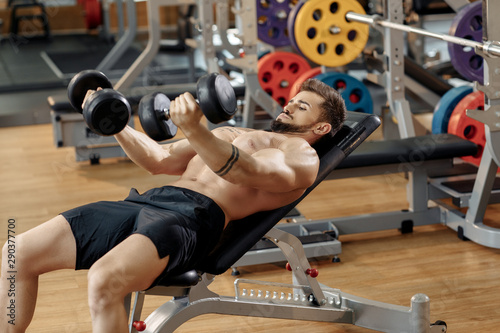 Muscular athletic young man bodybuilder fitness model exercise chest with dumbbell on the bench in gym. Top view.