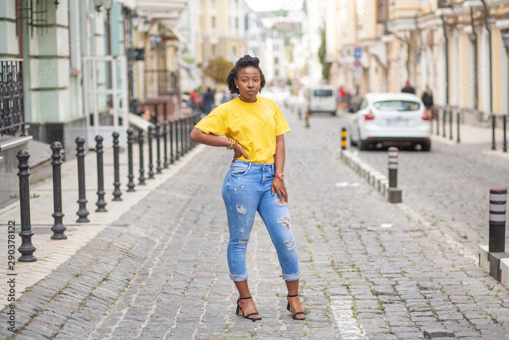 Lifestyle street girl. African American girl in a yellow T-shirt and blue  jeans stands on a city street. Stylish woman posing beautifully for a  photograph. She is very sweet and positive. Stock