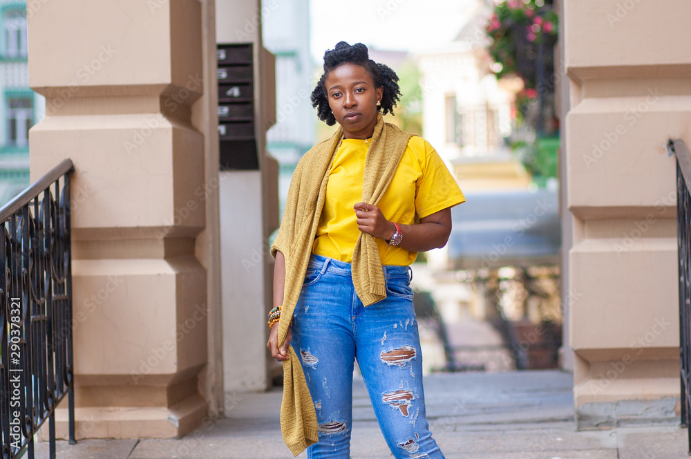 Lifestyle street girl. African American girl in a yellow T shirt and blue jeans stands on a city street. Stylish woman posing beautifully for a photograph. She is very sweet and positive. Stock