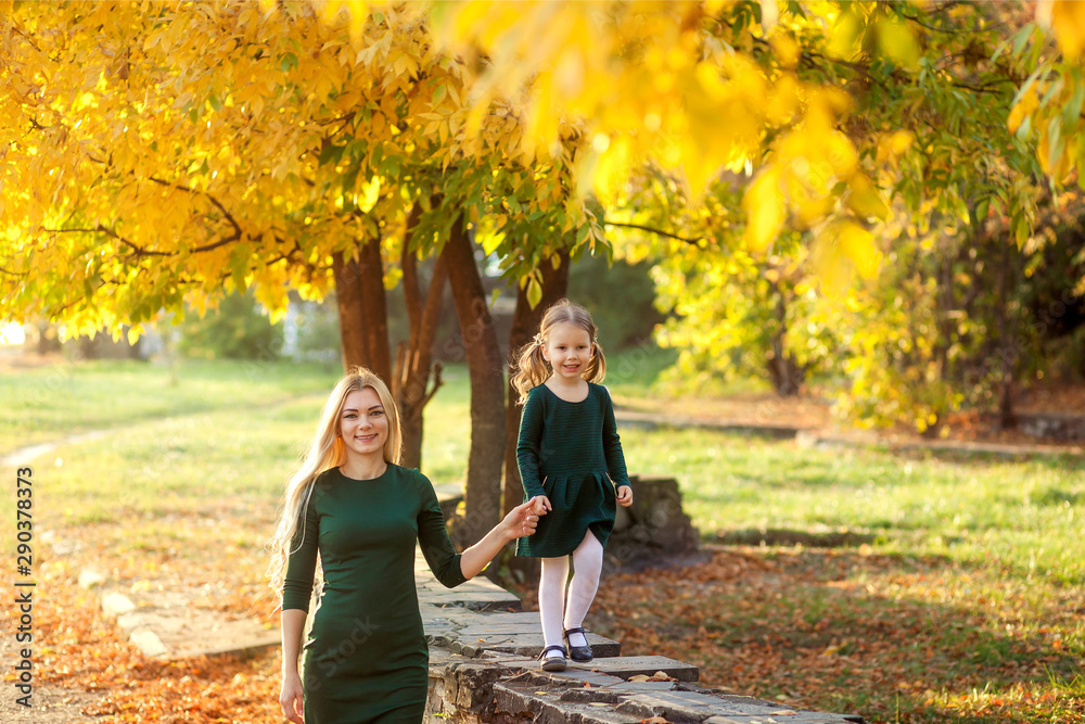 Fototapeta premium happy lovely mother and daughter for 3-4 years in identical green dresses in autumn in nature in the park, my mother has long blond hair, her daughter has wonderful curls, family walk