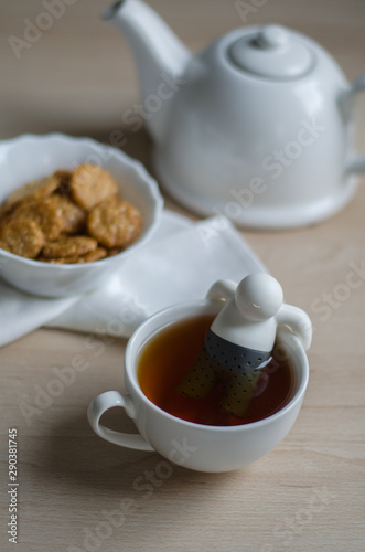 Cup of tea and biscuits with teapot on wooden background. Human figure in cup.