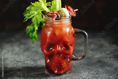 Bloody Mary cocktail in glass skull with celery sticks, pink salt, lime and canapes from canned vegetables. Halloween drink. 