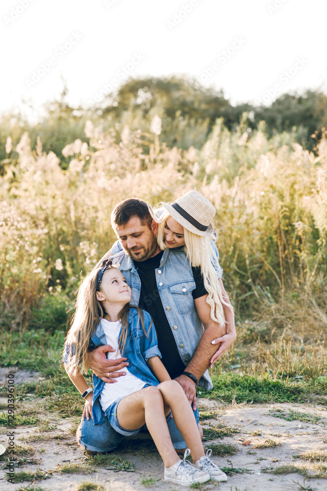 Happy young family, dad, mom and daughter, happily hugging, smiling and having a good time together. Young family resting on sunset background Happy young family, dad, mom and daughter, happily hugging, smiling and having a good time together. Young family resting on sunset background