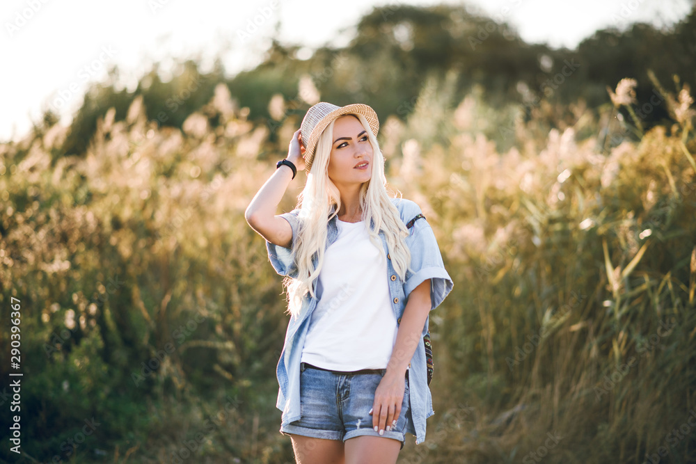 Beautiful girl with blond hair in a hat and shorts on a beautiful background with sunset. She poses and smiles.