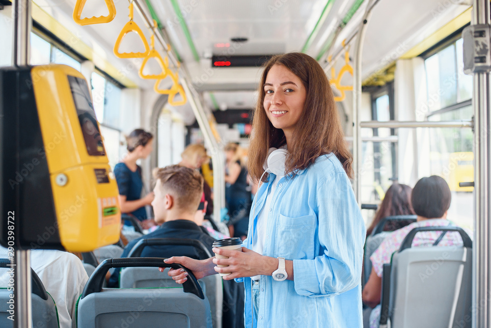Stylish woman in blue shirt enjoying trip in the modern tram or bus, stands with cup of coffee in the public transport. - obrazy, fototapety, plakaty 