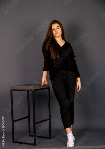 Young, beautiful girl in black clothes and white gym shoes.  A girl is standing in a beautiful pose by a high chair, studio shot on a plain background.