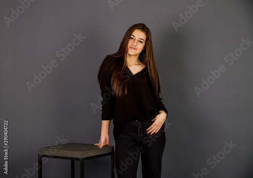 Young, beautiful girl in black clothes and white gym shoes.  A girl is standing in a beautiful pose by a high chair, studio shot on a plain background.