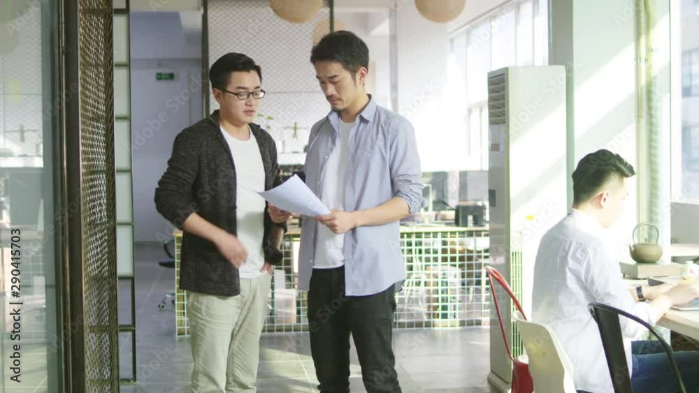two young asian business men discussing sales report whiling walking in office two young asian business men discussing sales report whiling walking in office