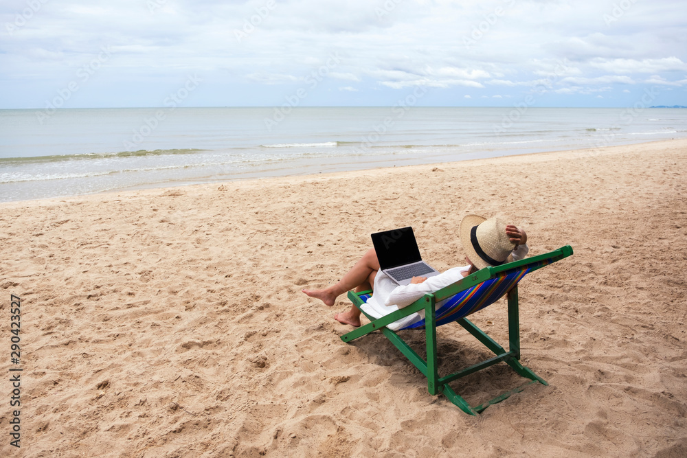 A woman using and typing on laptop computer while lying down on a beach chair A woman using and typing on laptop computer while lying down on a beach chair