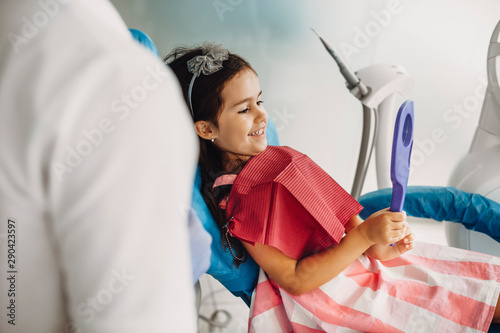 Lovely little girl looking in the mirror at her teeth after teeth surgery in a pediatric stomatology. Happy kid after teeth examination. photo