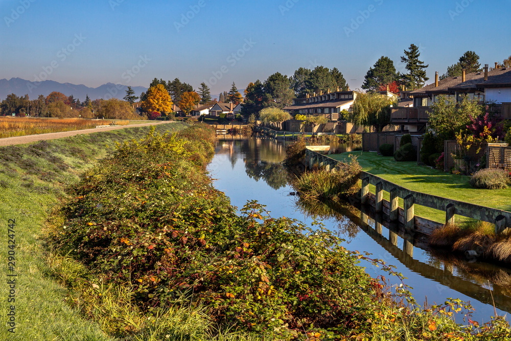 Residential area in a picturesque place on the banks of the canal with thickets of blackberry bushes on the other shore against the yellow autumn forest and mountain range on the horizon Residential area in a picturesque place on the banks of the canal with thickets of blackberry bushes on the other shore against the yellow autumn forest and mountain range on the horizon