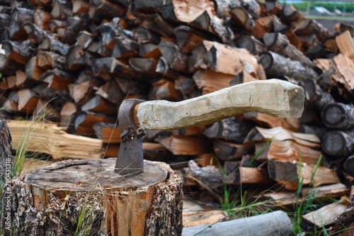 Old axe with wooden handle on stub. To cut and prepare firewood for winter. Woodpile with firewood at fence. Wood heating in village