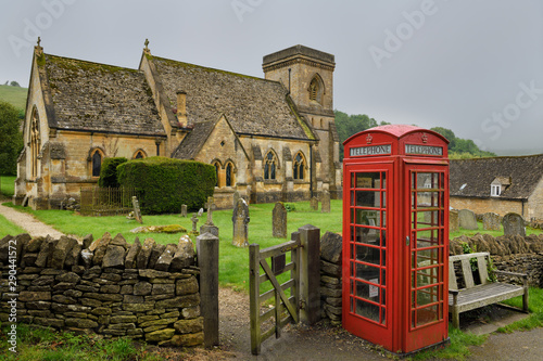 Red phone booth at 15th Century St Barnabas Anglican church with cemetery and stone wall gate in wet rainy weather in Snowshill Cotswold England photo