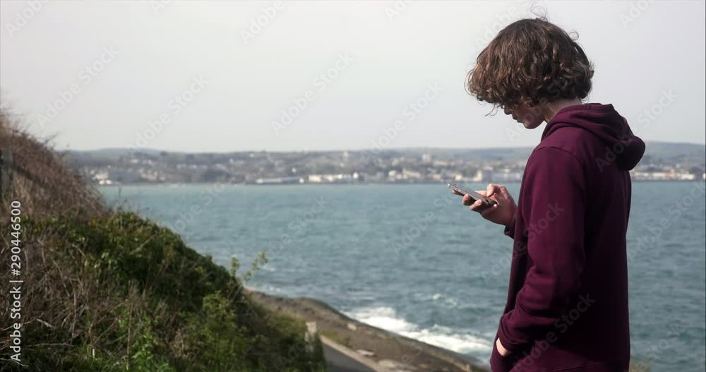 Young teenager boy searching social media on his mobile phone with coastal view in background, copy space. slow motion. Young teenager boy searching social media on his mobile phone with coastal view in background, copy space. slow motion.