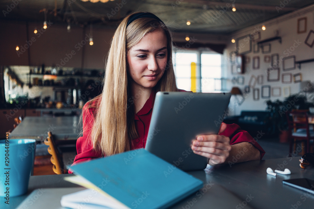Fototapeta premium Young woman in red suit reading tablet computer in big open space. Paperwork and cup of coffee on the table