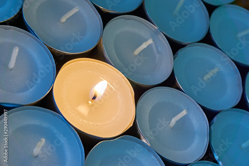 Closeup of tea lights with one being lit. A single warm orange flame between candles in blue shadow. Christmas, mourning, condolence, memorial, funeral or cremation ceremony concept. 