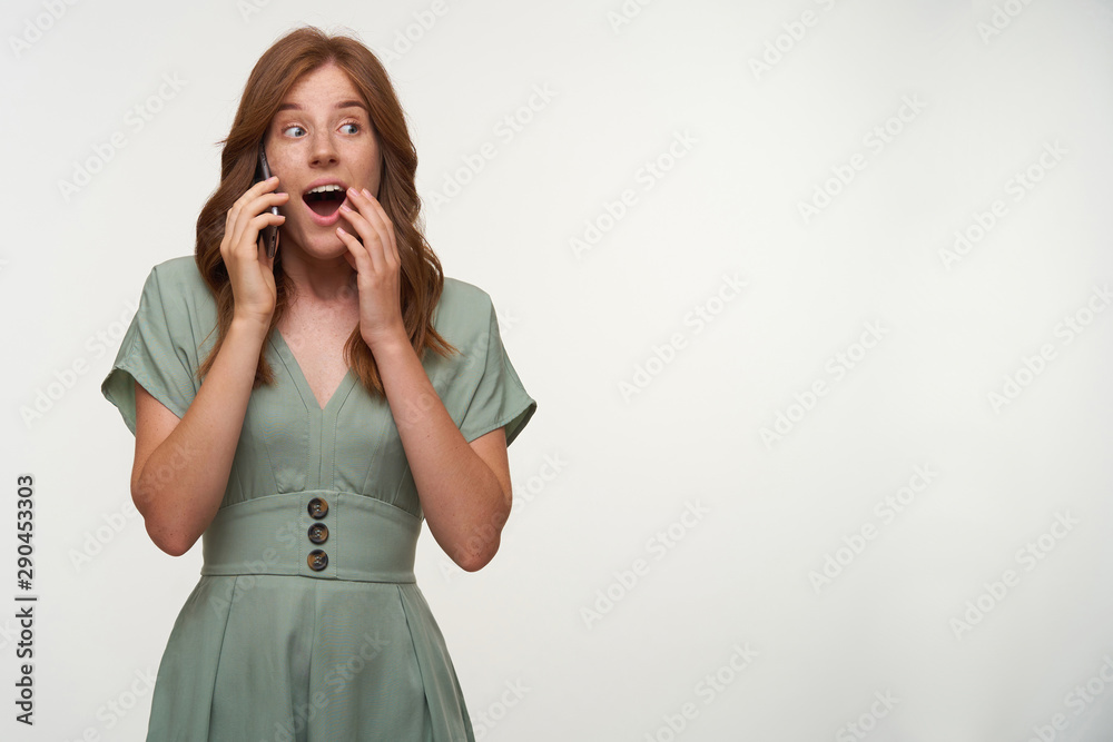 Amazed young beautiful female with red hair posing over white background, opening mouth widely and covering it with hand, holding smartphone by her ear Amazed young beautiful female with red hair posing over white background, opening mouth widely and covering it with hand, holding smartphone by her ear