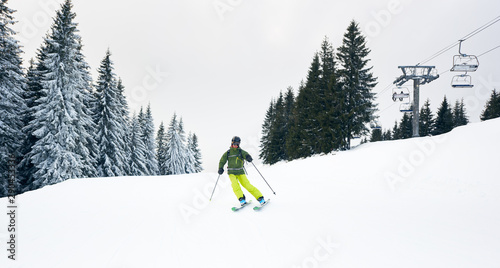 Male skier skiing on ski hillside at resort after taking chairlift on beautiful snow-covered mountain. Tourist backpacker freeriding on desert wooded slope. Ski chairlift and grey sky on background.