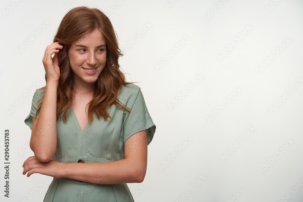 Indoor shot of beautiful redhead female with shy smile, looking aside and touching her hair, posing over white background in vintage dress Indoor shot of beautiful redhead female with shy smile, looking aside and touching her hair, posing over white background in vintage dress