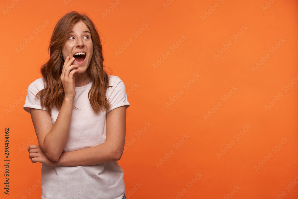 Cheerful good looking young female with red curly hair posing over orange background, touching her face with hand and opening mouth widely Cheerful good looking young female with red curly hair posing over orange background, touching her face with hand and opening mouth widely