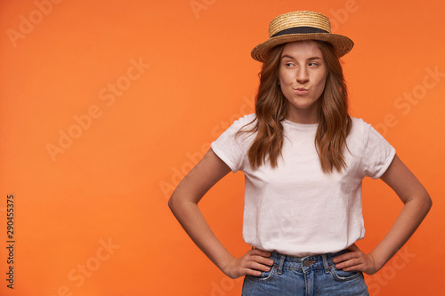 Portrait of pretty curly redhead woman standing over orange background with hands on waist, looking aside with cunning face, isolated over orange background