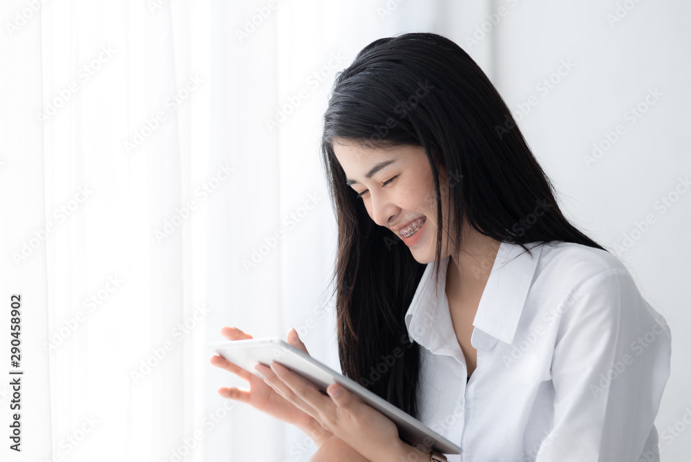 Beautiful Asian woman in white shirt using tablet near window in the morning. Beautiful Asian woman in white shirt using tablet near window in the morning.