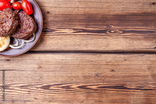 Tasty grilled burger meat with vegetables - onions and tomatoes - on brown vintage style plate on wooden background. Grilled food. Copy space.