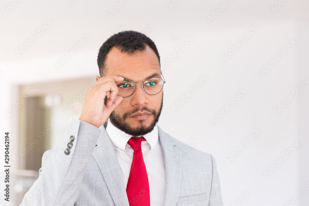 Serious business leader posing for camera in office hall. Businessman in office suit and tie adjusting eyeglasses and looking away. Glasses wearing concept Serious business leader posing for camera in office hall. Businessman in office suit and tie adjusting eyeglasses and looking away. Glasses wearing concept