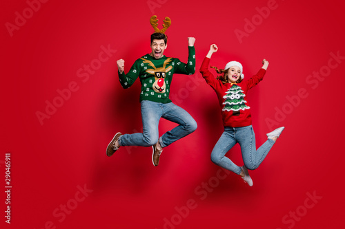 Full size photo of funky lady and guy jumping excited by x-mas prices wear ugly ornament jumpers and headwear isolated red color background
