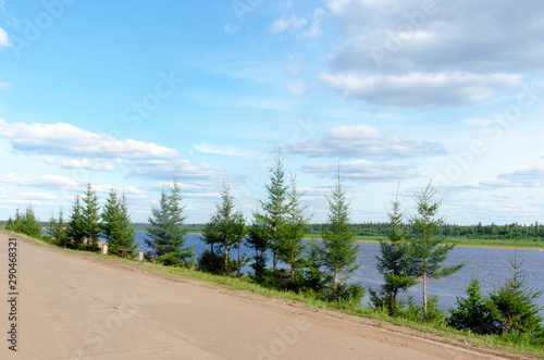 The road in the Northern Yakut village of ulus Suntar goes along the cliff above the vilyu river with small fir trees, a bench with urns under the clouds on a summer day.